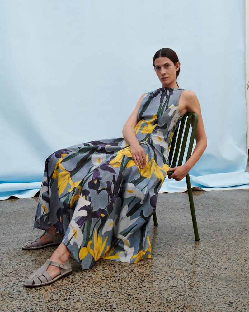 Woman wearing a floral print dress and beige sandals sitting leaning back in a green chair on a concrete surface in front of a draped white cloth