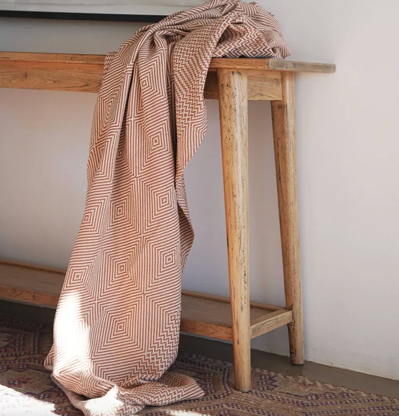 Patterned blanket draped over a wooden bench against a white wall.