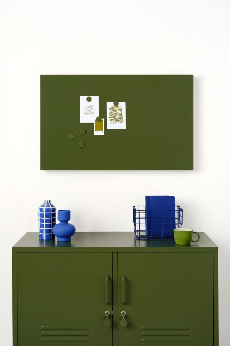 Green magnetic display board on a white wall above a green locker cabinet with decorative items.