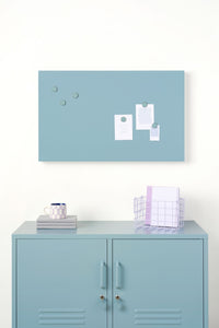 Light blue magnetic display board with white magnets on a white wall above a light blue locker.