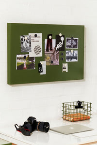 Green magnetic board with photos and notes held by magnets on a white wall above a desk with a camera and laptop.