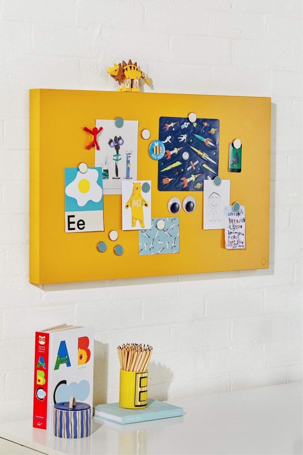 Yellow magnetic board with various magnets and a small figurine on a white wall.