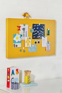 Yellow magnetic board with various magnets and a small figurine on a white wall.