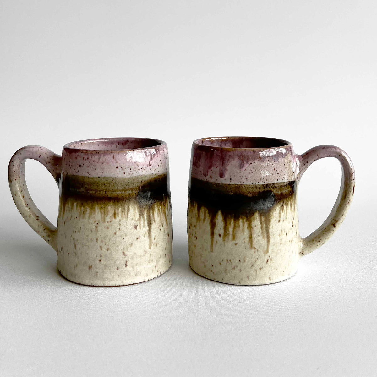 Two ceramic mugs with light and dark neutral and pink glaze details on a white background