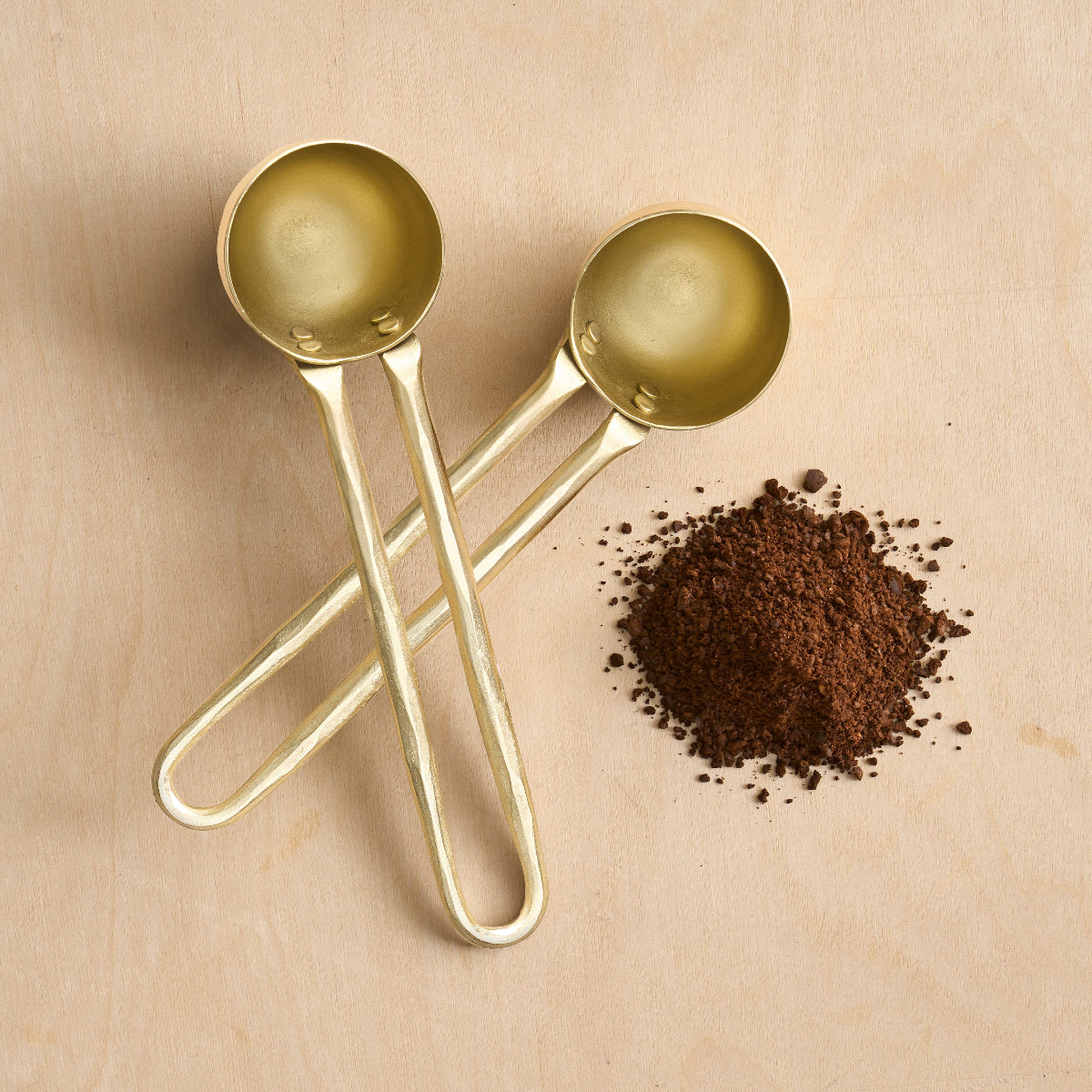 Two brass coffee spoons nest to coffee grounds on a wooden surface