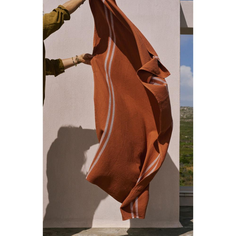 Rust color bath towel with white stripes draped by a person against a neutral background