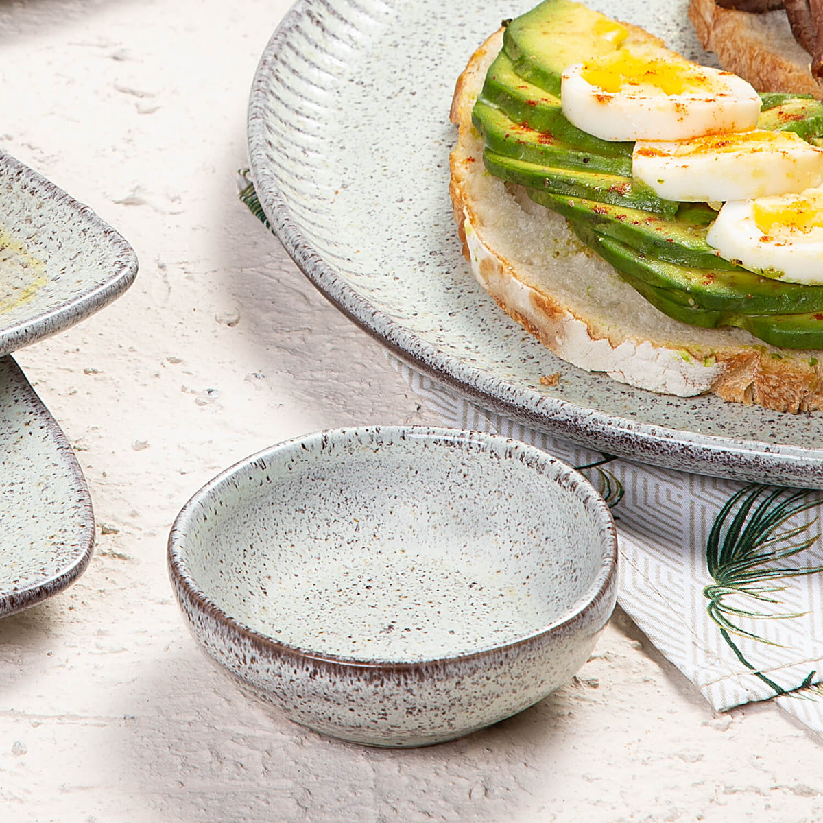 Speckled ceramic ramekin with a plate of avocado toast on a light surface.
