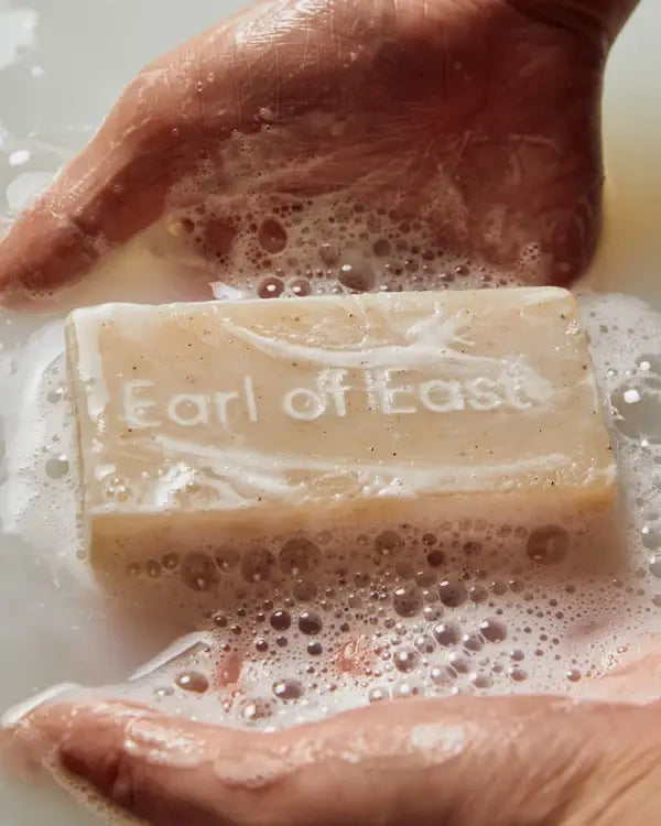 to hands reaching into soapy water surrounding a beige bar of soap with the words 'Earl of East' imprinted on the bar 