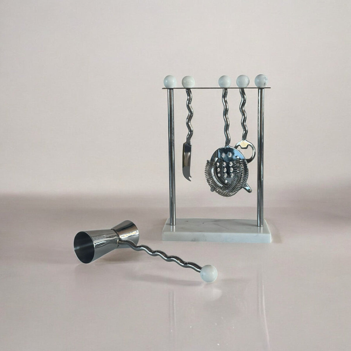 Cocktail tool set with jigger, bar spoon, and strainer on a stand against a light background