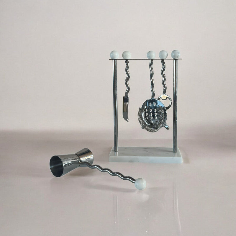 Cocktail tool set with jigger, bar spoon, and strainer on a stand against a light background