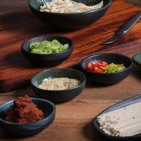 Assorted condiments in small black ramekins on a wooden surface