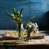 Two glass vases with greenery and white flowers on a textured tablecloth against a blue wall.