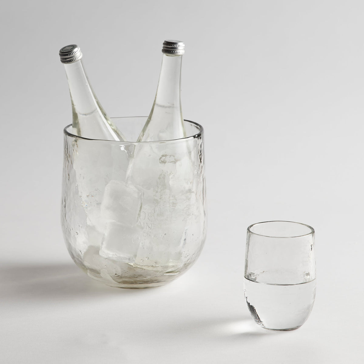 Clear glass ice bucket with two bottles and a glass on a light gray background