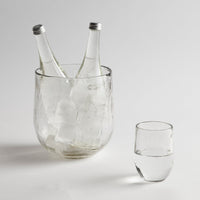 Clear glass ice bucket with two bottles and a glass on a light gray background