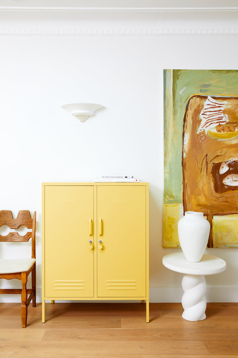 Yellow cabinet next to a white chair and a white sculpture on a wooden floor with a colorful abstract painting on the wall.