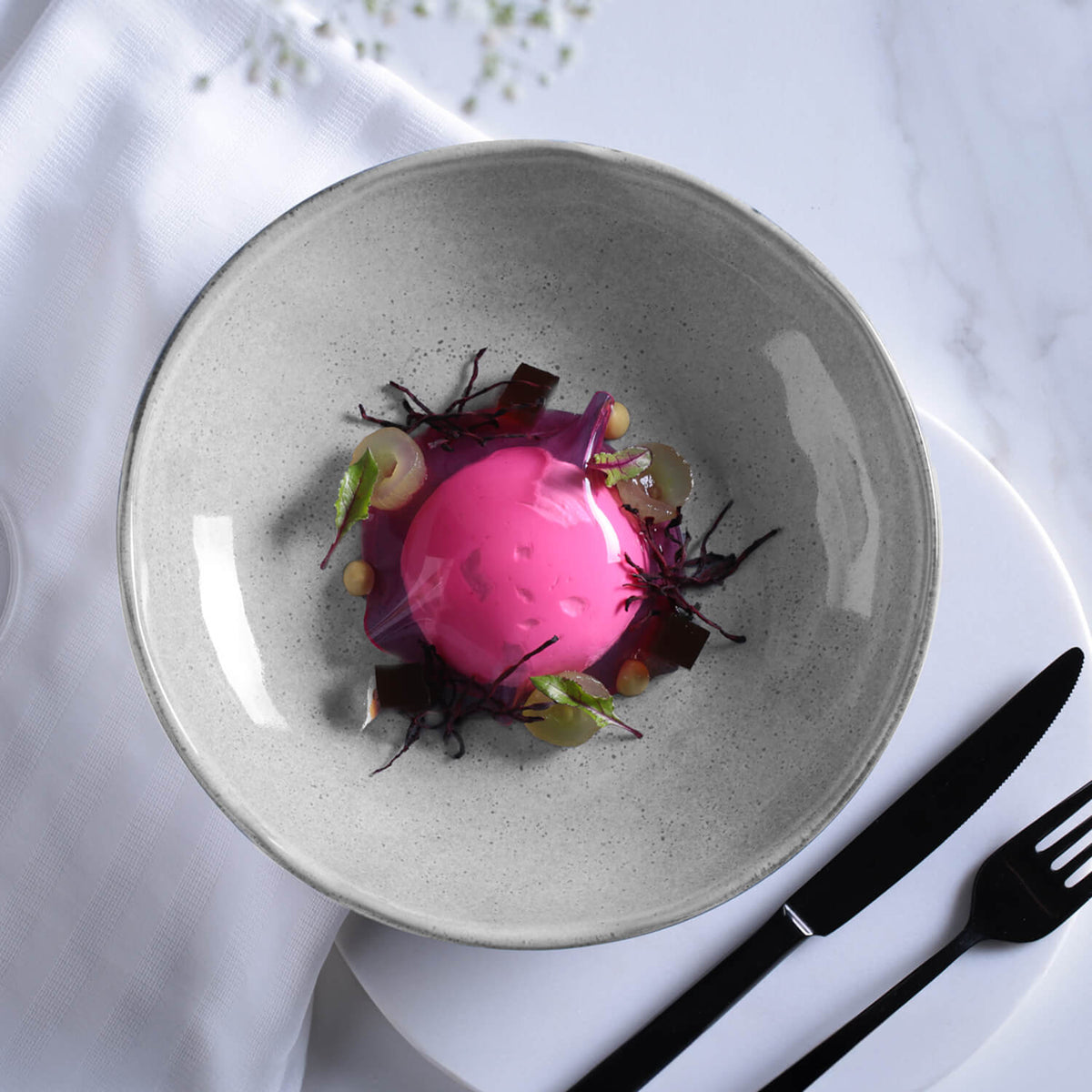 Pink dessert on a gray plate with garnishes, served on a white tablecloth with cutlery.