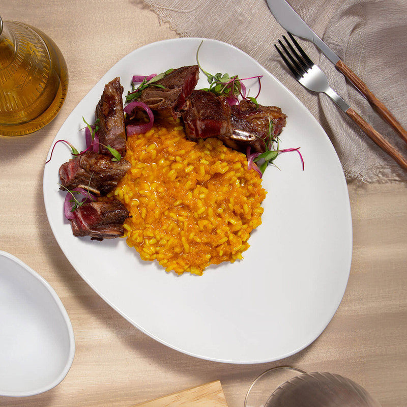 White plate of yellow risotto with grilled meat and vegetables on a wooden table.