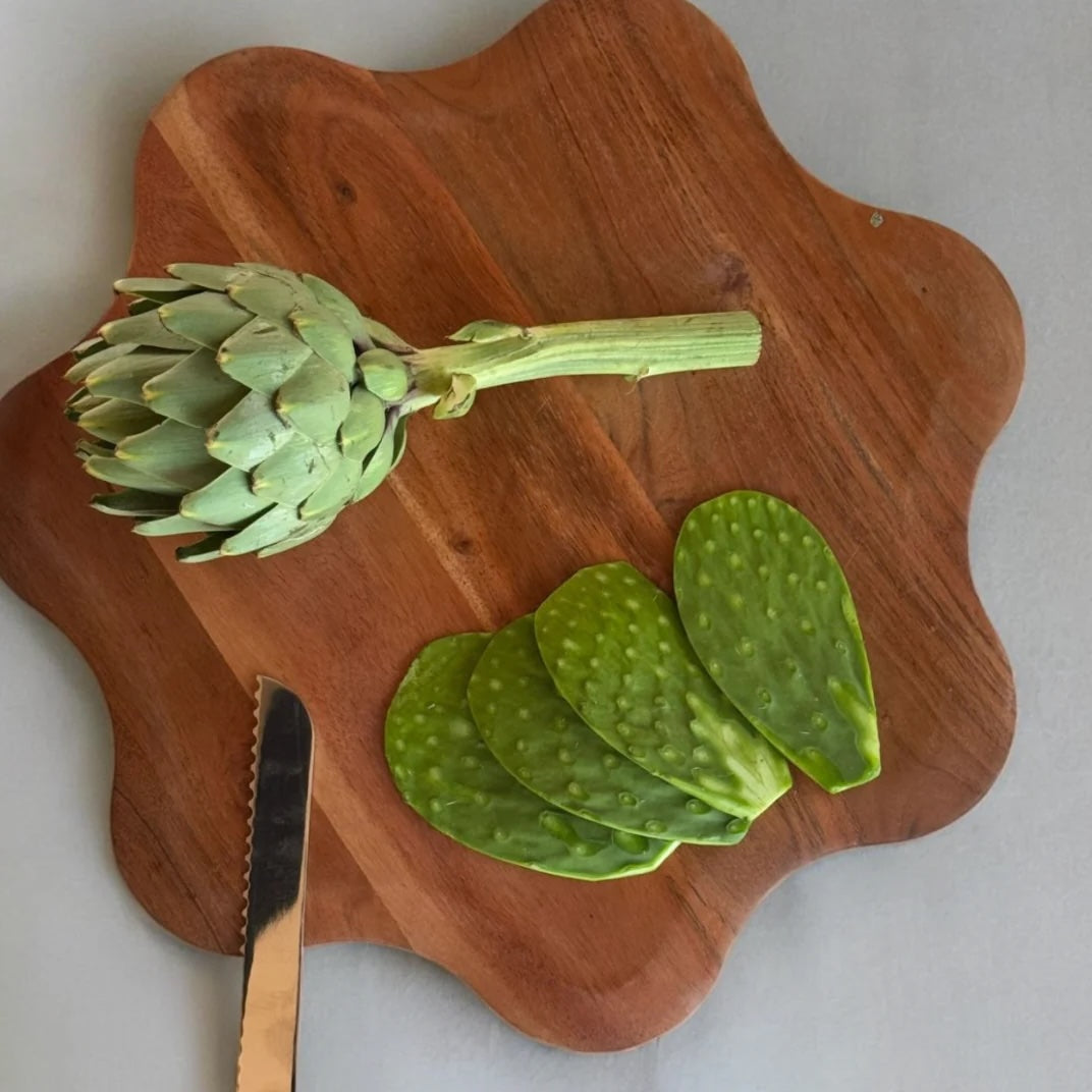 Artichoke and cactus paddles on a wooden cutting board with a knife.