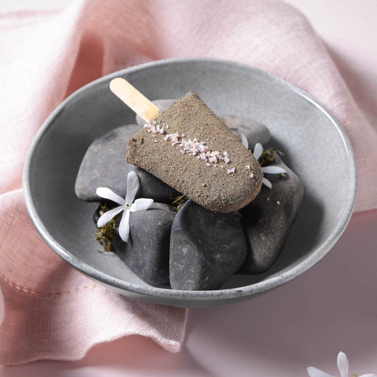 Dark chocolate popsicle with pink sprinkles on a bed of stones in a gray ceramic bowl, against a pink fabric background.