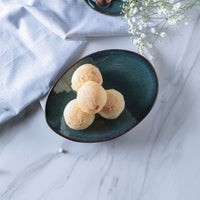 Three round pastries in a dark green ceramic bowl on a marble surface with a light blue cloth and white flowers in the background.