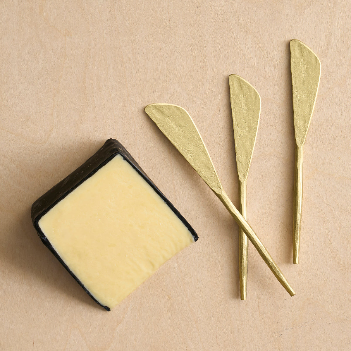 Three brass spreader knives and a slice of cheese on a wooden surface