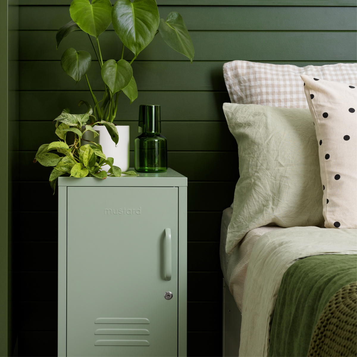 Green locker style nightstand with plant and water carafe next to a bed with pillows against a green wall.