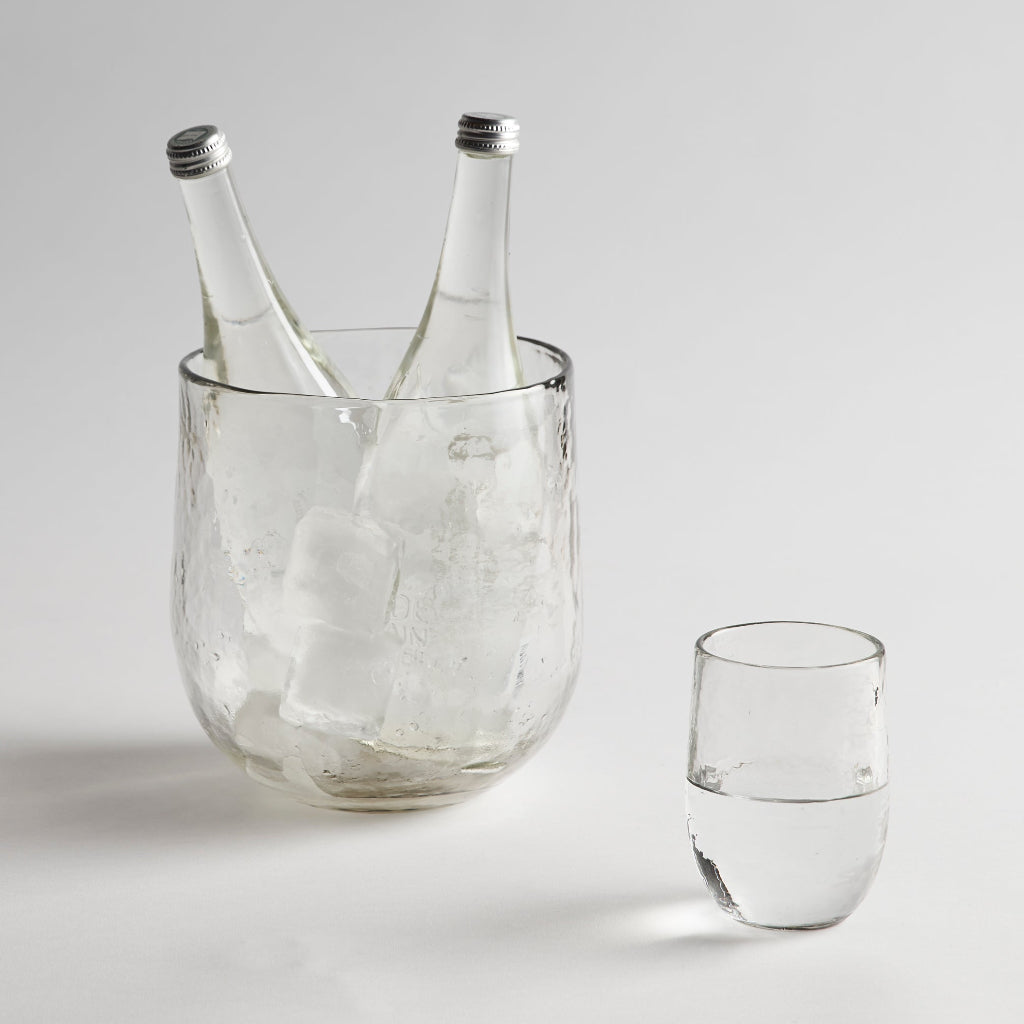 Glass ice bucket filled with two bottles and a drinking glass of water on a white background