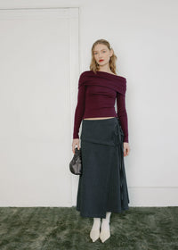 A model stands against a white wall wearing a dark burgundy off-the-shoulder long-sleeve top and a dark gray pleated wrap skirt, holding a small black handbag.