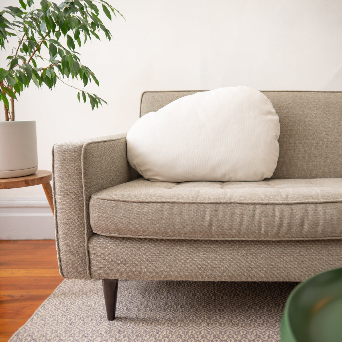 Beige sofa with a white pillow in a room with a plant and wooden floor.