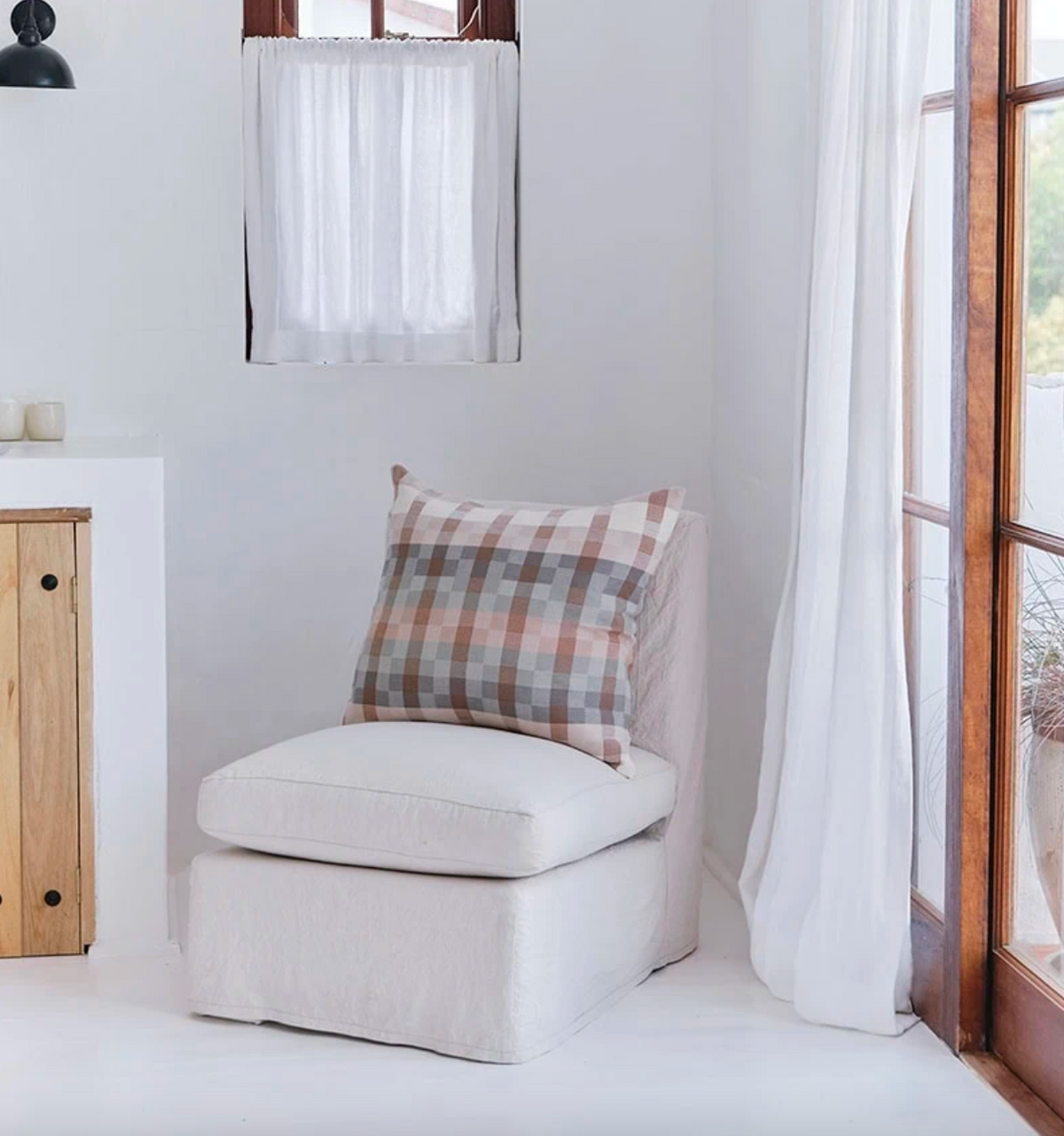 White armchair with a plaid pillow in a bright room with wooden furniture and large windows.