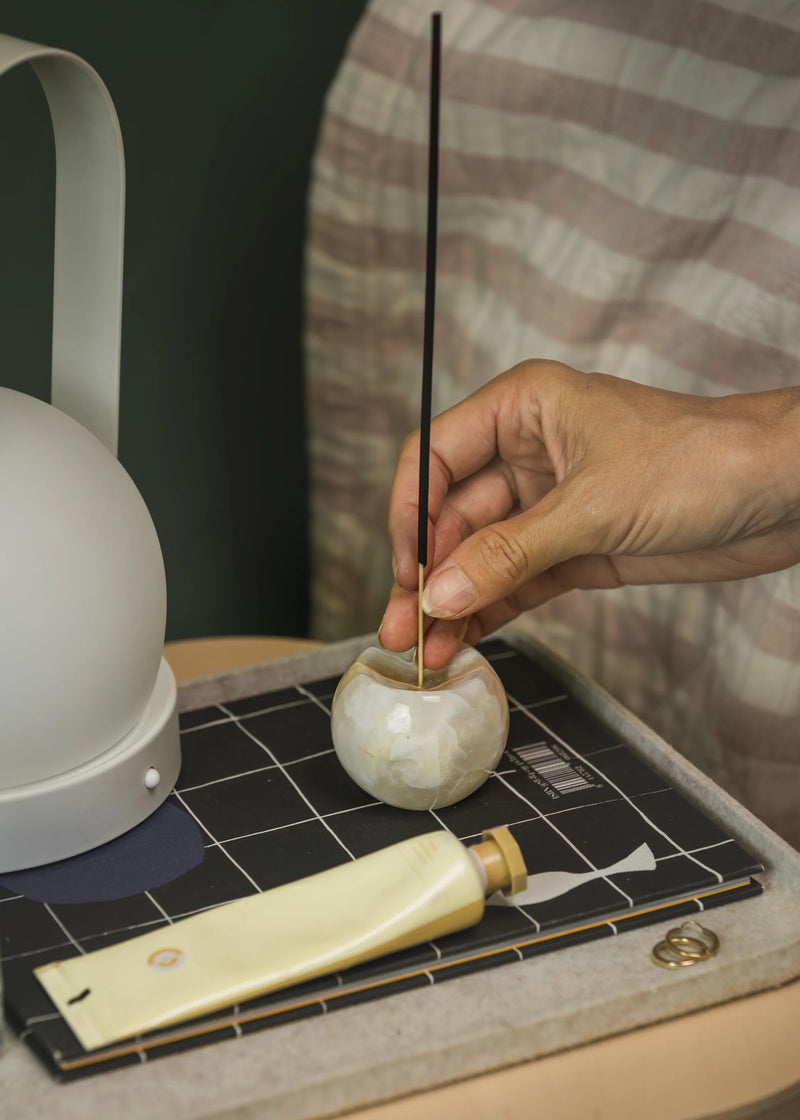 Hand placing an incense stick into a small stone holder on a checkered surface.