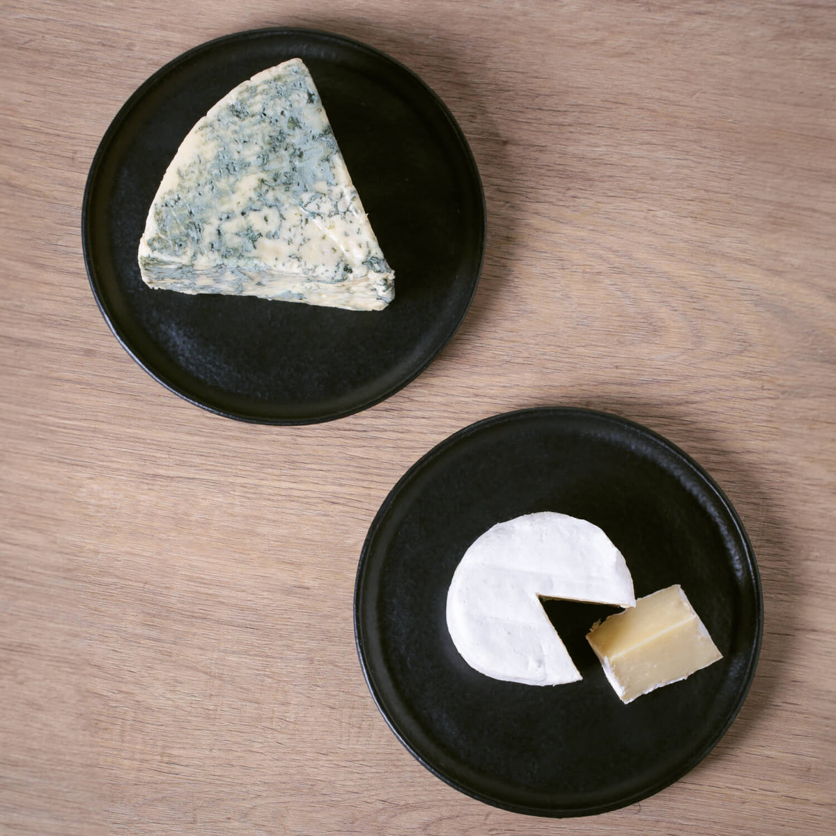 Two black plates with different types of cheese on a wooden surface