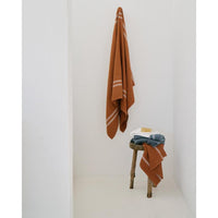 Rust color towel hanging on a white wall with other towels draped over a wooden stool.