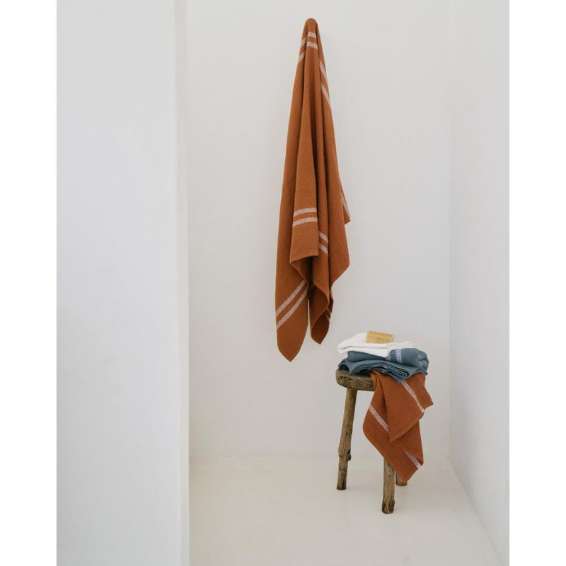 Rust color towel hanging on a white wall with other towels draped over a wooden stool.
