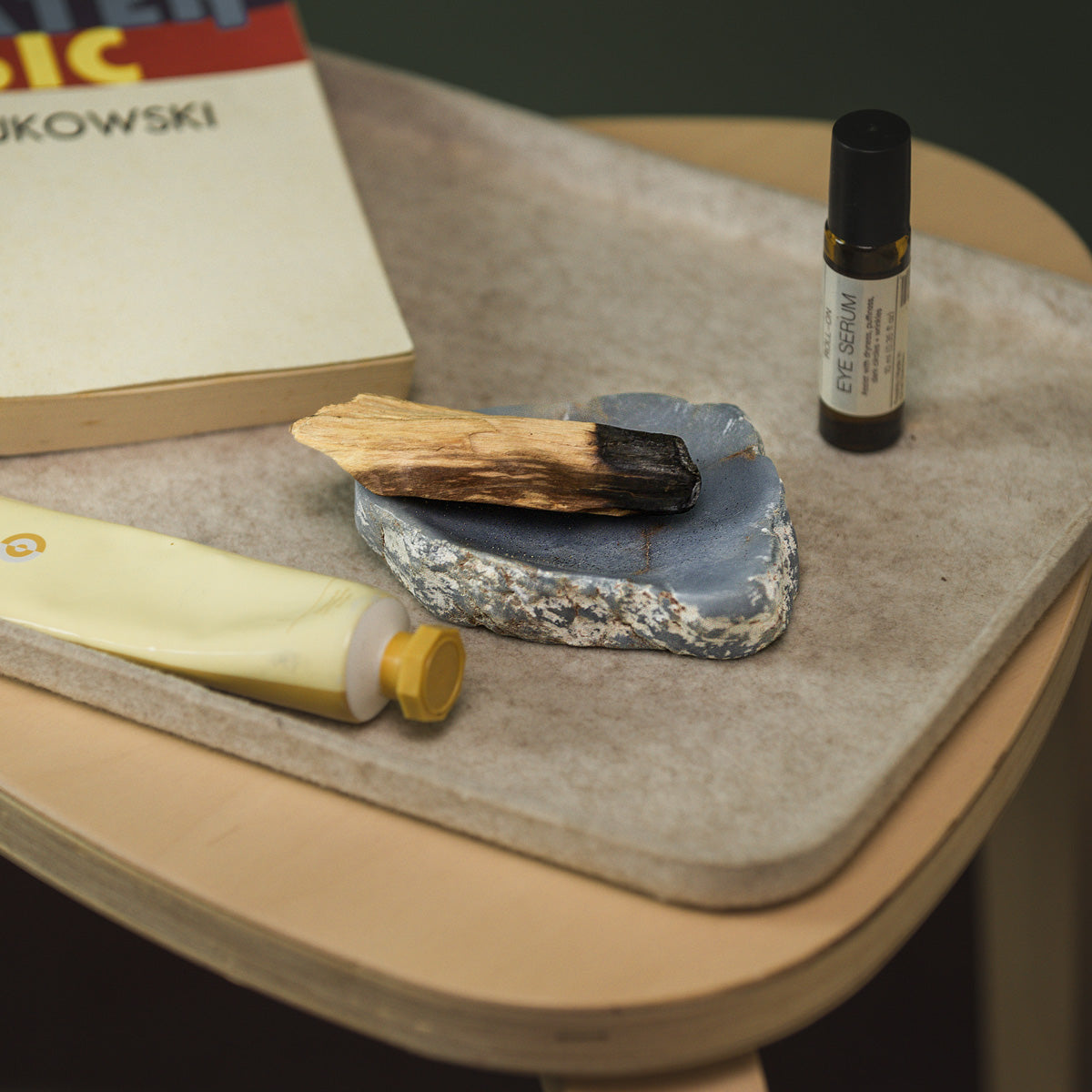Small bottle of eye serum, palo Santo stick on a stone dish, and tube of hand cream on a stone surface with a book in the background.