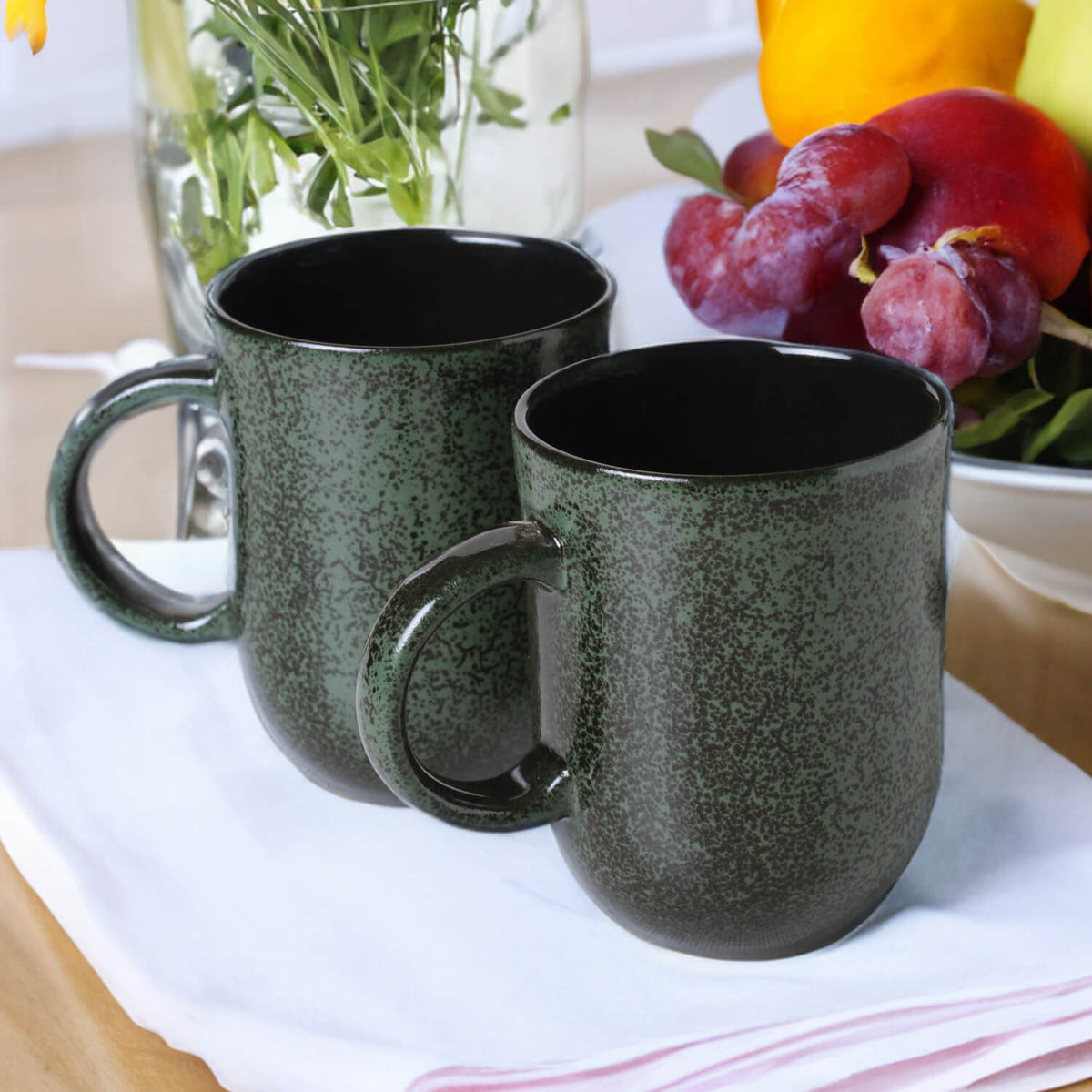 Two green speckled mugs on a table with fruits and a vase in the background.