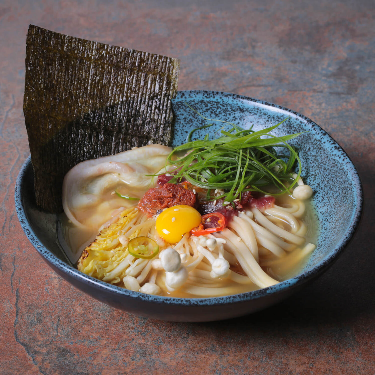 Noodle dish with vegetables and a poached egg in a blue bowl on a textured surface