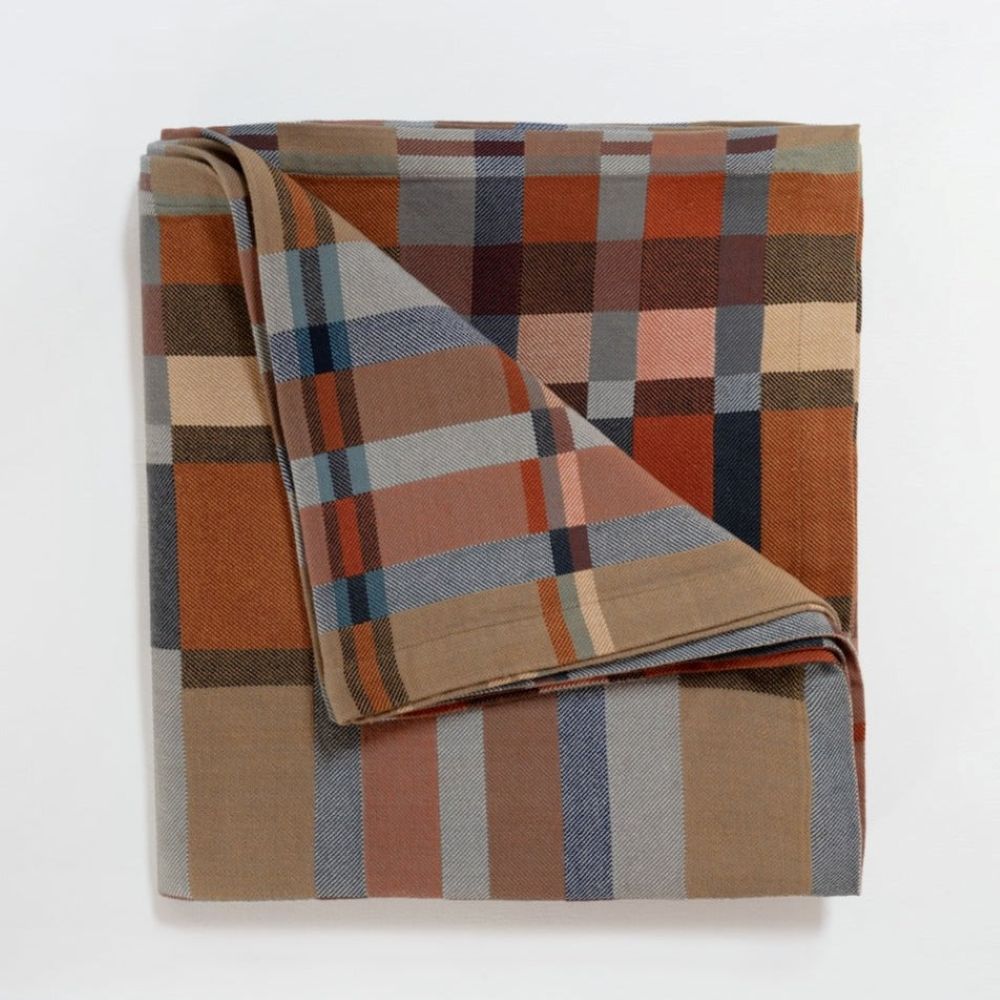 A folded plaid fabric item, likely a blanket or throw, featuring a warm autumn color palette of rust orange, brown, beige, and muted blue-gray on a white background.