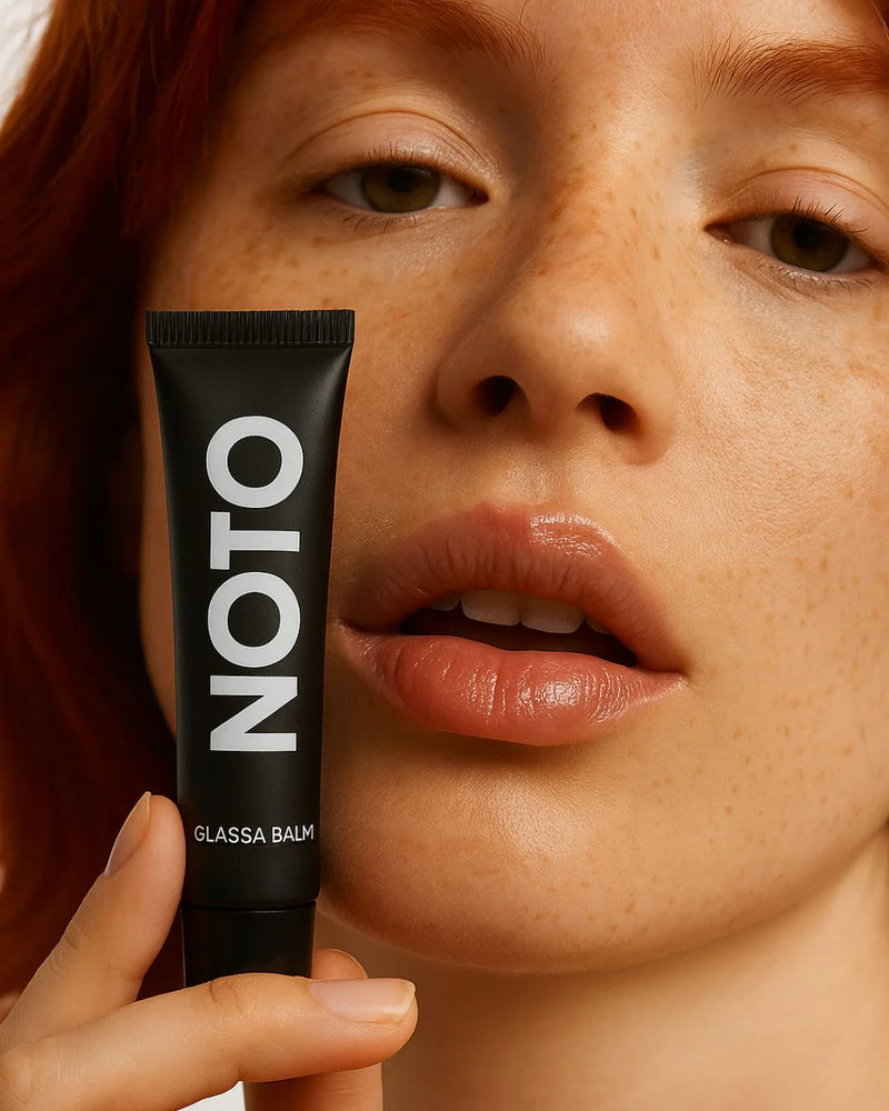 Person holding a black tube labeled 'Noto' Glassa Balm near their lips against a neutral background