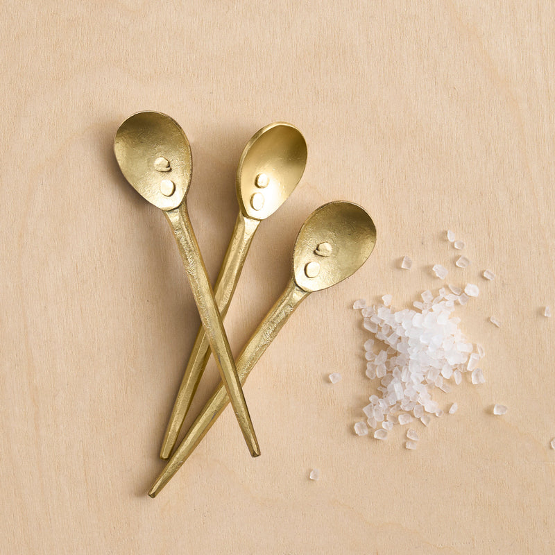 Three mini brass spoons next to salt on a wooden surface