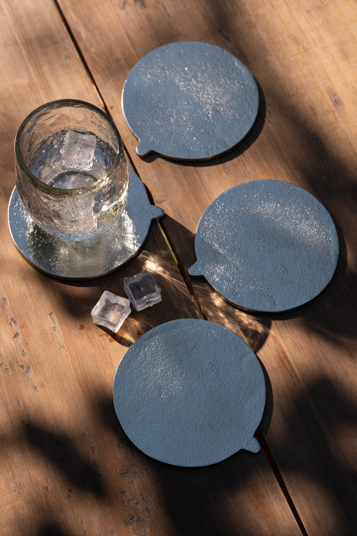 Set of silver coasters on a wooden surface with a glass of water and ice cubes.