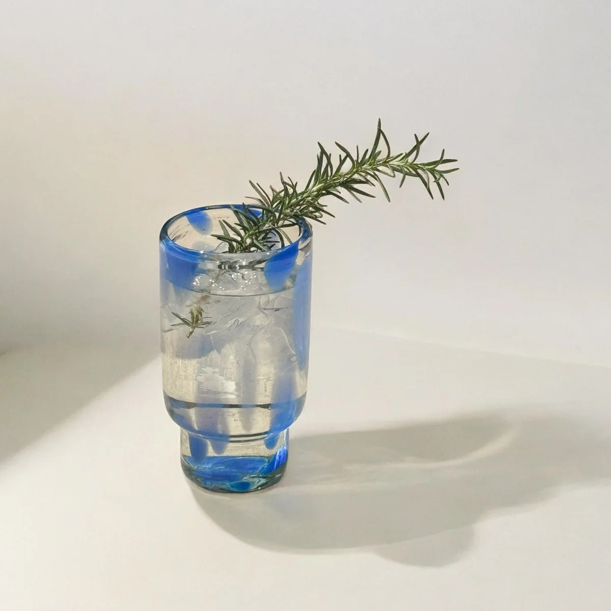 Clear glass with blue pattern containing a drink and a sprig of rosemary on a white background