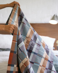 Person holding a plaid blanket over a bed with a wooden headboard.