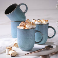 Four blue mugs, two filled with toasted marshmallows on a white surface.