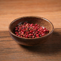 Brown ceramic ramekin filled with red peppercorns on a wooden surface