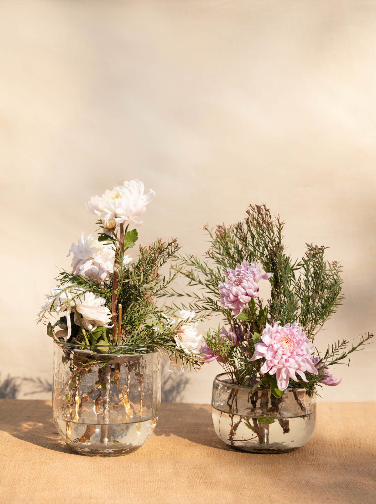 Two glass vases with floral arrangements on a beige surface and background