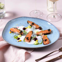 Plated blue dish of salmon with garnishes on a light pink tablecloth with wine glasses in the background.