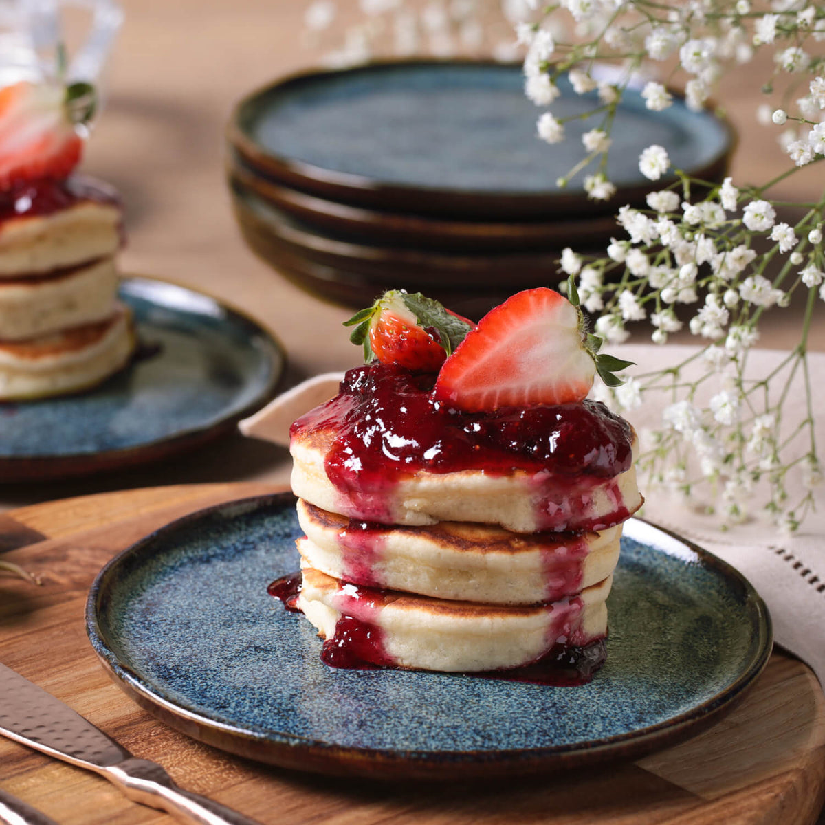 Stack of pancakes with strawberry sauce on a blue plate, surrounded by flowers and additional plates.