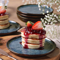 Stack of pancakes with strawberry sauce on a blue plate, surrounded by flowers and additional plates.