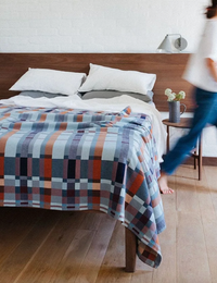 A bed in a modern bedroom with a colorful geometric plaid blanket draped over it, alongside a person walking past on the right.